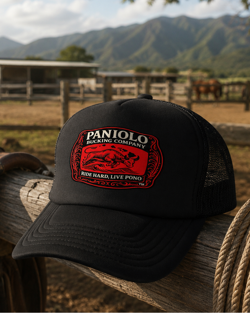 A black trucker hat with a red and white embroidered 'Paniolo Racing Company' logo, resting on a wooden fence post in a rustic outdoor barn setting with mountains in the background.