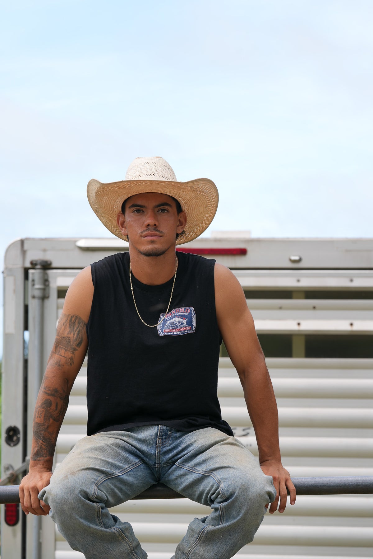 A man wearing a straw cowboy hat, a black sleeveless shirt, and light blue jeans sits on a metal rail in front of a white horse trailer under a pale blue sky.