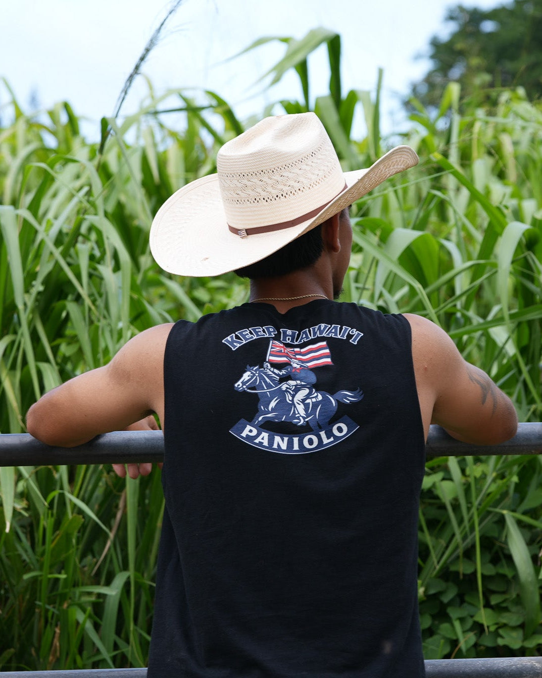 A man seen from behind wearing a black tank top with a detailed graphic on the back, a beige cowboy hat, and leaning on a metal railing in front of a dense green field.