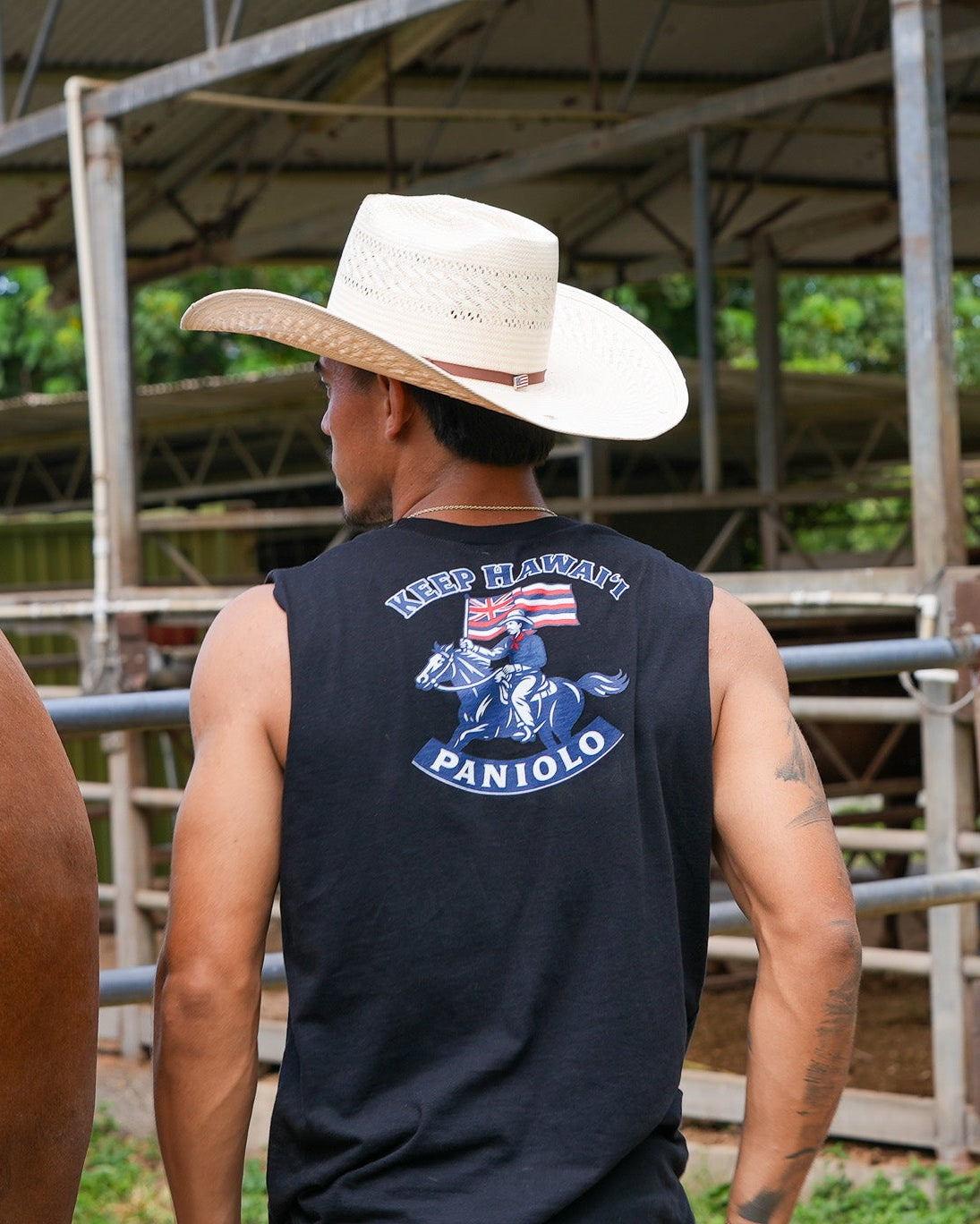 A man wearing a white cowboy hat and a black sleeveless shirt with a 'Paniolo' ranch graphic, standing in a barn next to a brown horse.